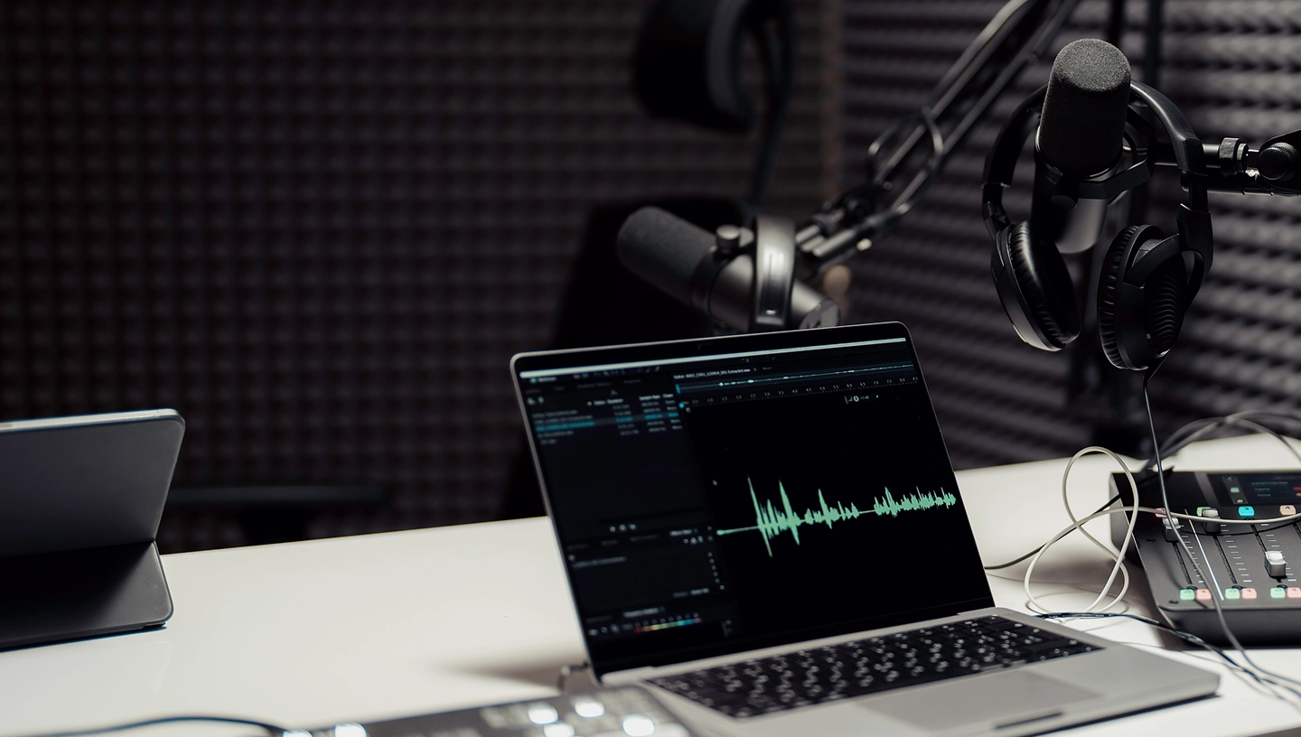 Professional podcast studio setup with laptop displaying audio waveform editing software, studio microphone, headphones, and audio mixer on white desk with dark acoustic foam background